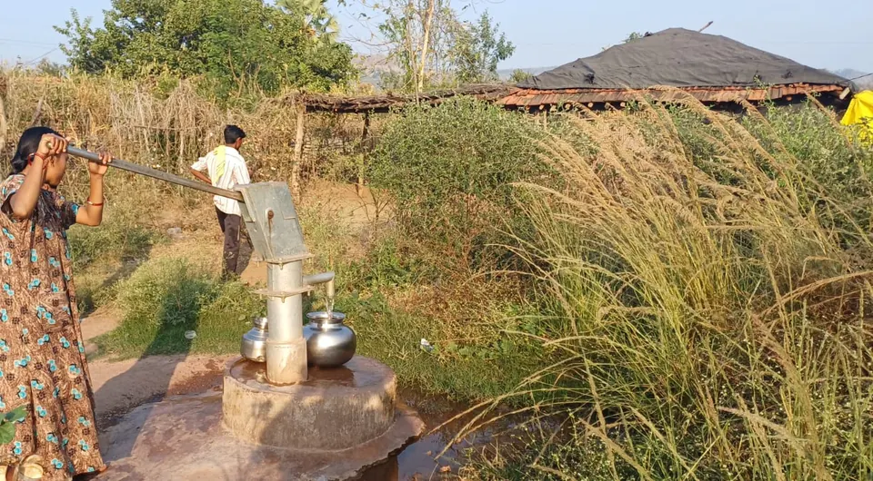 Woman pumping water from a village borewell hand pump into steel pots